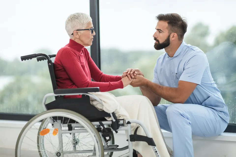 Support worker kneeling beside a participant in a wheelchair, holding hands and talking